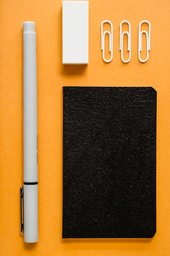 Flat lay image of office supplies on an orange background. Includes pen, notebook, paperclips, and eraser.
